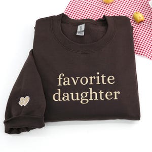 A dark chocolate sweatshirt featuring “favorite daughter” embroidery in soft beige thread.
The sleeve is accented with a small double-heart detail stitched in matching tones.