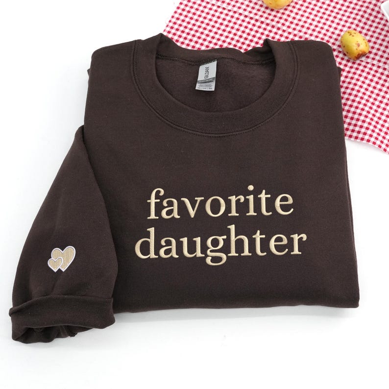 A dark chocolate sweatshirt featuring “favorite daughter” embroidery in soft beige thread.
The sleeve is accented with a small double-heart detail stitched in matching tones.
