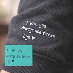 May include: Dark gray sweatshirt sleeve with white embroidered text: "I love you Always and forever, Lyn" with a heart symbol. A teal sticky note repeats the same message. The sweatshirt is made of a soft, textured fabric.