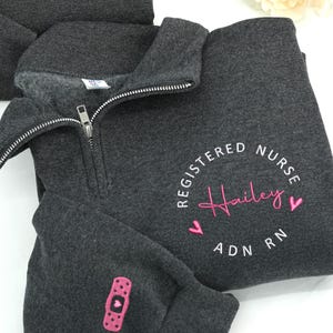 May include: Dark grey quarter-zip sweatshirt with a white zip. The sweatshirt features the text "REGISTERED NURSE" in a semi-circle, with the name "Hailey" in pink script. Below the name are the letters "ADN RN" and two pink hearts. A pink bandage graphic is on the sleeve.