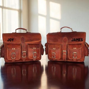 May include: Two brown leather satchels with multiple front pockets and brass buckles. Each bag has a top handle and a flap closure. One bag is labeled "JOY" and the other "JAMES". The bags are sitting on a dark brown surface.