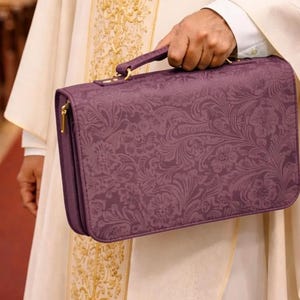 Może przedstawiać: Prostokątna fioletowa teczka z teksturowanym wzorem kwiatowym i górnym uchwytem, trzymana przy białej szacie liturgicznej ze złotym haftowanym krzyżem. Teczka posiada zamek błyskawiczny w kolorze złotym wzdłuż bocznych krawędzi.