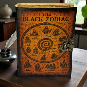 May include: A leather-bound journal titled "The Black Zodiac" with a detailed illustration of zodiac symbols. The cover is dark brown with gold accents and a metal clasp. The journal is displayed on a wooden table.