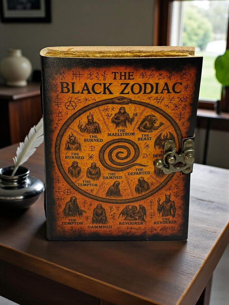 May include: A leather-bound journal titled "The Black Zodiac" with a detailed illustration of zodiac symbols. The cover is dark brown with gold accents and a metal clasp. The journal is displayed on a wooden table.