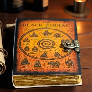 May include: A leather-bound journal titled "The Black Zodiac" with an aged, antique appearance. The cover displays zodiac illustrations and is fastened with a metal clasp. The pages are yellowed, giving the impression of an old, possibly magical, book.