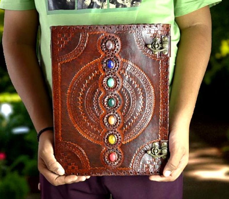 May include: A brown leather journal with a decorative embossed design and a series of colorful gemstones. The journal has a clasp closure and is held open. The gemstones are red, yellow, green, blue, and purple.