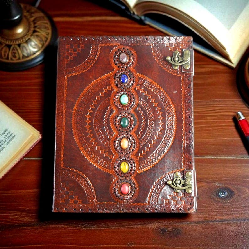 May include: A brown leather journal with a decorative embossed design and a series of colorful gemstones. The journal has a metal clasp and is open on a wooden surface. The cover features a chakra design.