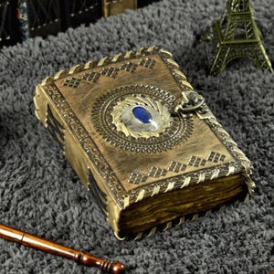 Könnte beinhalten: Ein Tagebuch im Vintage-Stil mit einem messingfarbenen Einband und aufwendigen Details. Der Einband zeigt einen zentralen blauen Stein, der von einem verzierten Design umgeben ist. Das Tagebuch wird mit einem Metallverschluss geschlossen und hat gealterte Seiten.