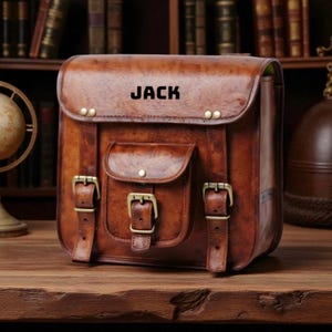 May include: A brown leather satchel with the name "JACK" embossed on the flap. The bag features a front pocket, brass buckles, and straps. The satchel is sitting on a wooden surface.