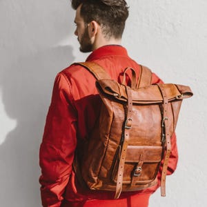Könnte beinhalten: Ein brauner Lederrucksack mit Rolltop-Verschluss, zwei vertikalen Schnallenriemen, einer Vordertasche mit Einzelschnallenverschluss und verstellbaren Schultergurten. Die Tasche hat eine strukturierte Oberfläche und einen Tragegriff oben.