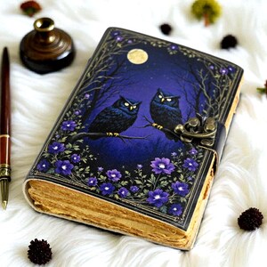 May include: A leather-bound journal with a night sky design featuring two owls perched on a branch. The cover is a deep blue with a full moon and purple flowers. The journal has a metal clasp and aged paper.