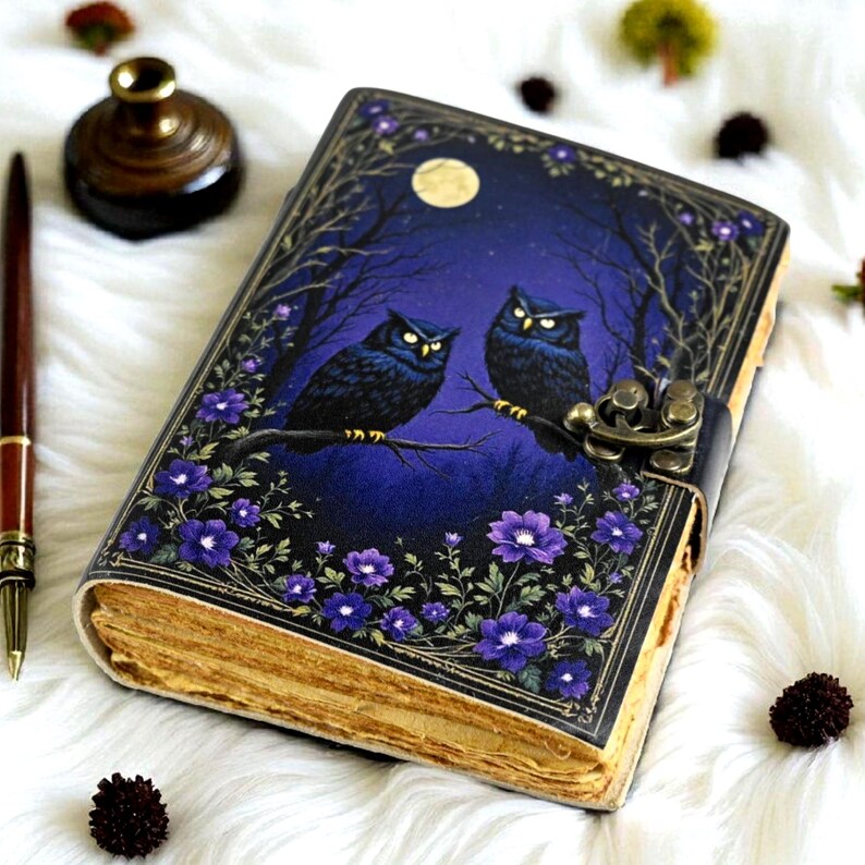 May include: A leather-bound journal with a night sky design featuring two owls perched on a branch. The cover is a deep blue with a full moon and purple flowers. The journal has a metal clasp and aged paper.