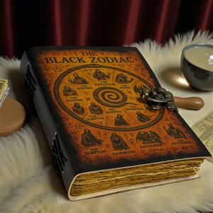 May include: A leather-bound journal titled "THE BLACK ZODIAC" with an illustrated cover featuring zodiac symbols. The journal has a clasp closure and aged, yellowed pages. The cover is brown and black.