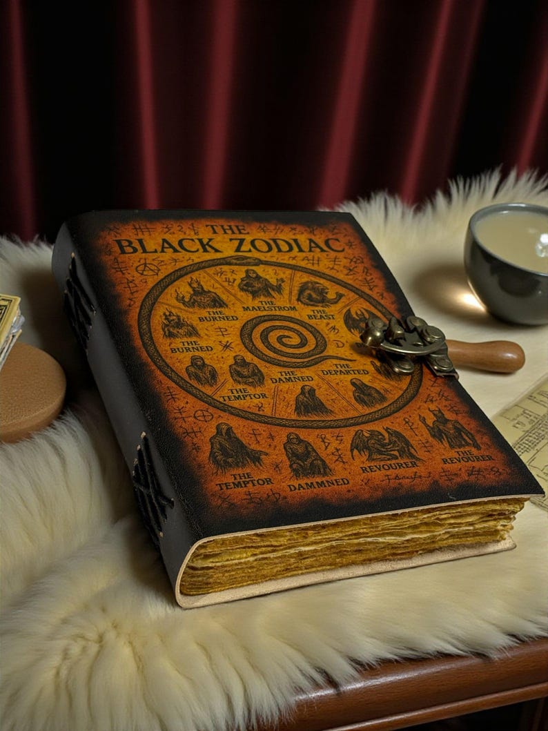 May include: A leather-bound journal titled "THE BLACK ZODIAC" with an illustrated cover featuring zodiac symbols. The journal has a clasp closure and aged, yellowed pages. The cover is brown and black.