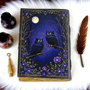 May include: A decorative journal with a dark blue cover featuring two owls perched on a branch under a full moon. The cover is framed with purple flowers and branches. A brown feather and ink bottles are next to the journal.