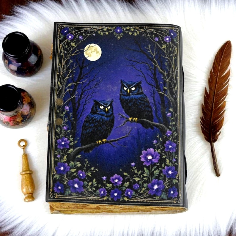 May include: A decorative journal with a dark blue cover featuring two owls perched on a branch under a full moon. The cover is framed with purple flowers and branches. A brown feather and ink bottles are next to the journal.