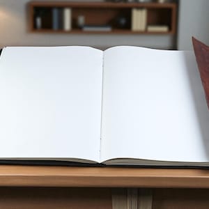 May include: An open, blank journal with a dark cover and a brown leather spine. The journal is lying on a wooden surface, with a lamp and chair visible in the background. The pages are white and ready for writing.