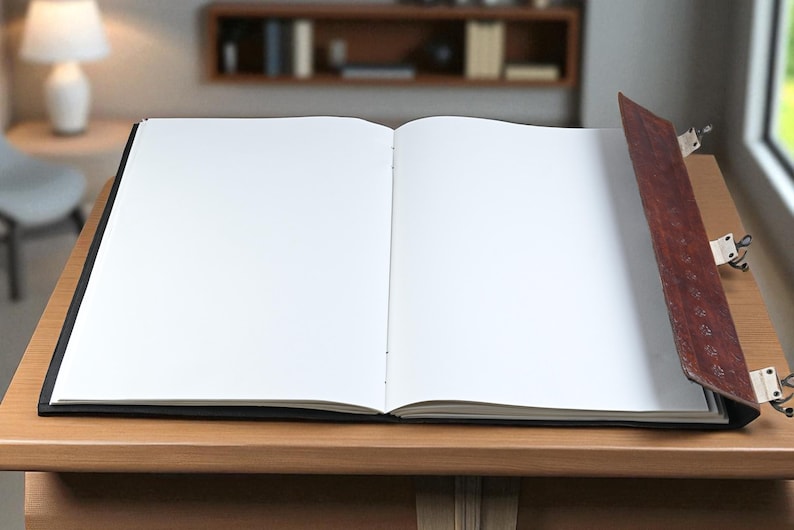 May include: An open, blank journal with a dark cover and a brown leather spine. The journal is lying on a wooden surface, with a lamp and chair visible in the background. The pages are white and ready for writing.