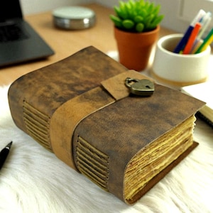 May include: A vintage-style leather journal with a heart-shaped lock. The journal has a distressed brown cover and a tan leather strap closure. The pages are aged, and the journal is on a white surface.