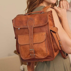 Könnte beinhalten: Ein hellbrauner Lederrucksack mit einem Tragegriff, einer großen Vordertasche mit Schnallenverschluss und Seitentaschen mit Metallbeschlägen. Der Rucksack verfügt über sichtbare Nähte an den Kanten und verstellbare Schultergurte.