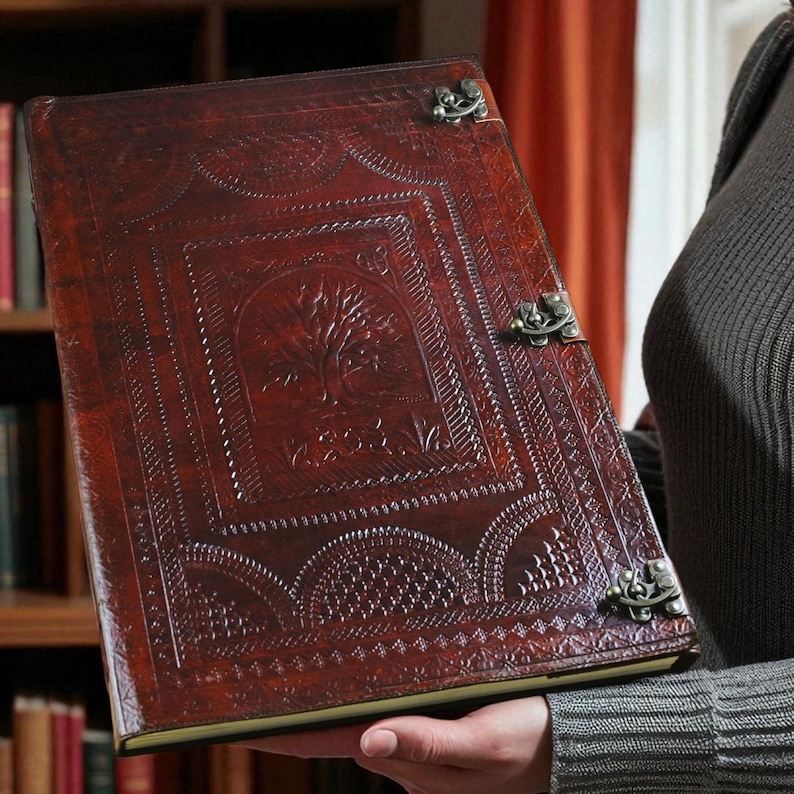 May include: A brown leather journal with an embossed tree design on the cover. The journal has decorative metal clasps and intricate border patterns. The cover is a rich, reddish-brown color, and the pages are bound with a gold edge.