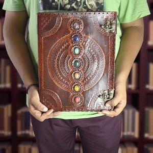 May include: A brown leather journal with an embossed design and colorful gemstones. The journal has a decorative clasp and is held in front of a blurred library background. The journal's design features a central circular pattern with seven gemstones.