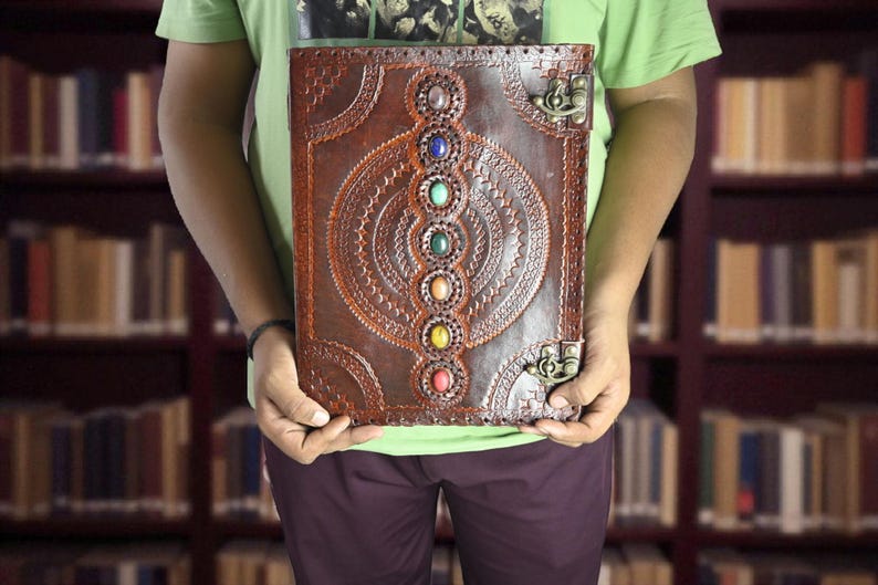 May include: A brown leather journal with an embossed design and colorful gemstones. The journal has a decorative clasp and is held in front of a blurred library background. The journal's design features a central circular pattern with seven gemstones.