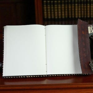 May include: An open, blank journal with a dark brown leather cover and a decorative clasp. A vintage inkwell with a feather quill and a small oil lamp sit nearby on a wooden surface, suggesting a writing or study theme.