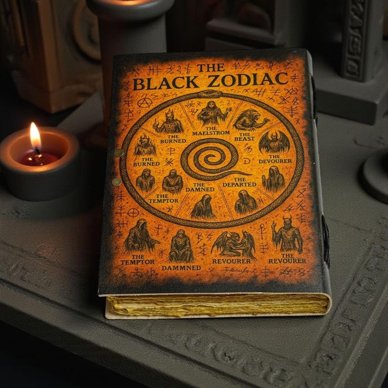 May include: A hardback book titled "THE BLACK ZODIAC" with gold-edged pages. The cover displays a circular illustration of zodiac figures and symbols in black and gold. The book is on a dark grey surface, with a lit candle in the background.