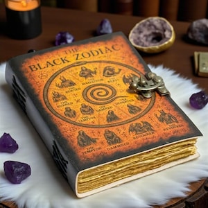 May include: A leather-bound book titled "The Black Zodiac" with an orange cover featuring zodiac symbols and the text. The book has a metal clasp and aged, yellowed pages. The book is on a white fur-like surface.