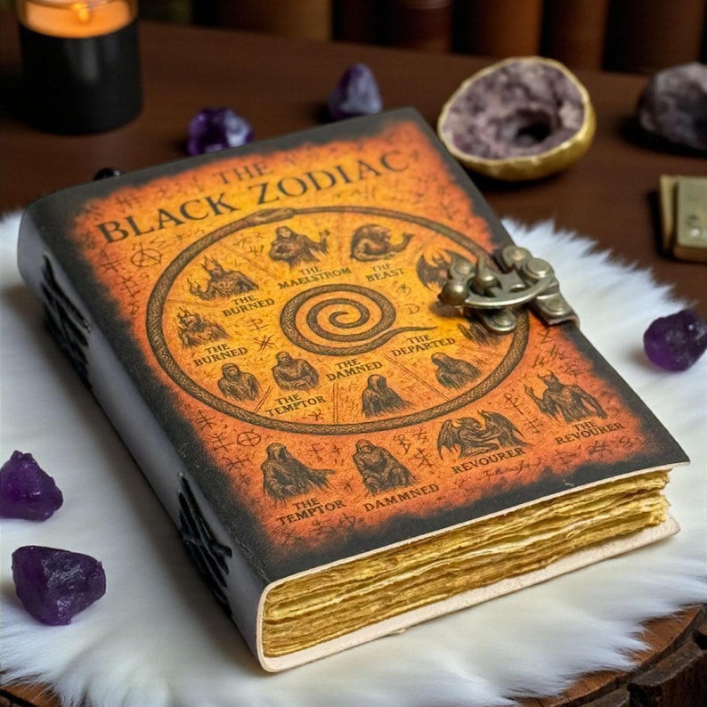 May include: A leather-bound book titled "The Black Zodiac" with an orange cover featuring zodiac symbols and the text. The book has a metal clasp and aged, yellowed pages. The book is on a white fur-like surface.