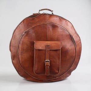 May include: A large, circular, brown leather bag with a handle and a small front pocket. The bag features a zip closure and a buckle on the pocket. The leather has a distressed appearance.