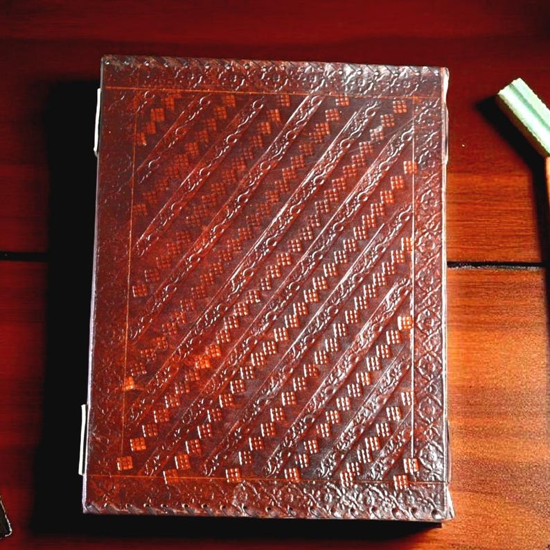 May include: A rectangular, brown leather-bound journal with a detailed embossed pattern. The cover features diagonal lines and a decorative border. The journal is closed, with the pages visible on the left side.