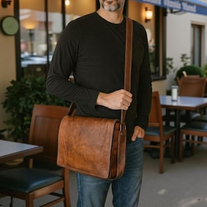 Op de afbeelding: Een bruine leren messenger bag met een schouderriem. De tas heeft een rechthoekige vorm met een klepsluiting. Het leer heeft een distressed look. De persoon draagt een zwart shirt met lange mouwen en een blauwe jeans.