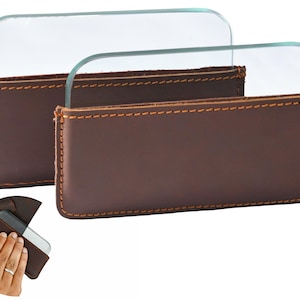 May include: Two clear glass lenses are displayed in brown leather cases with orange stitching. The cases are rectangular with rounded edges. The lenses are curved and rectangular. The image shows a close-up of the lenses and cases.