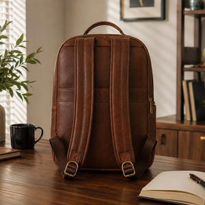 K&ouml;nnte beinhalten: Ein brauner Lederrucksack mit zwei gepolsterten Schultergurten mit verstellbaren Metallschnallen, einem Tragegriff an der Oberseite und einer vertikalen Rei&szlig;verschlusstasche an der Seite. Das Design hat eine glatte, einfarbige Oberfl&auml;che.