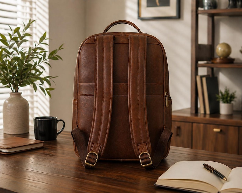 K&ouml;nnte beinhalten: Ein brauner Lederrucksack mit zwei gepolsterten Schultergurten mit verstellbaren Metallschnallen, einem Tragegriff an der Oberseite und einer vertikalen Rei&szlig;verschlusstasche an der Seite. Das Design hat eine glatte, einfarbige Oberfl&auml;che.