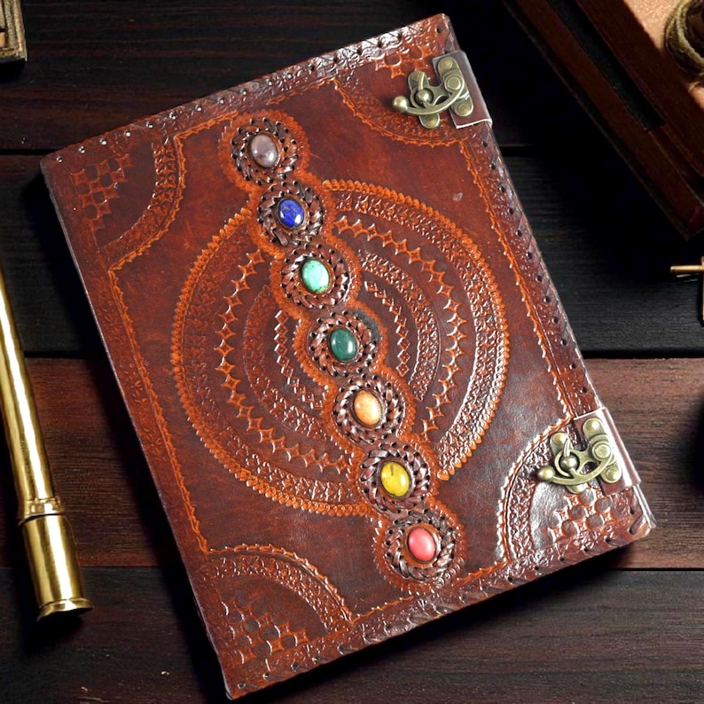 May include: A brown leather journal with a decorative embossed design and a series of colorful gemstones. The journal features a metal clasp closure and intricate circular patterns.