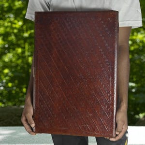 May include: A large, rectangular, dark brown leather-bound book with a textured, diagonal pattern. The book is held by a person, with a blurred green background of trees and foliage.