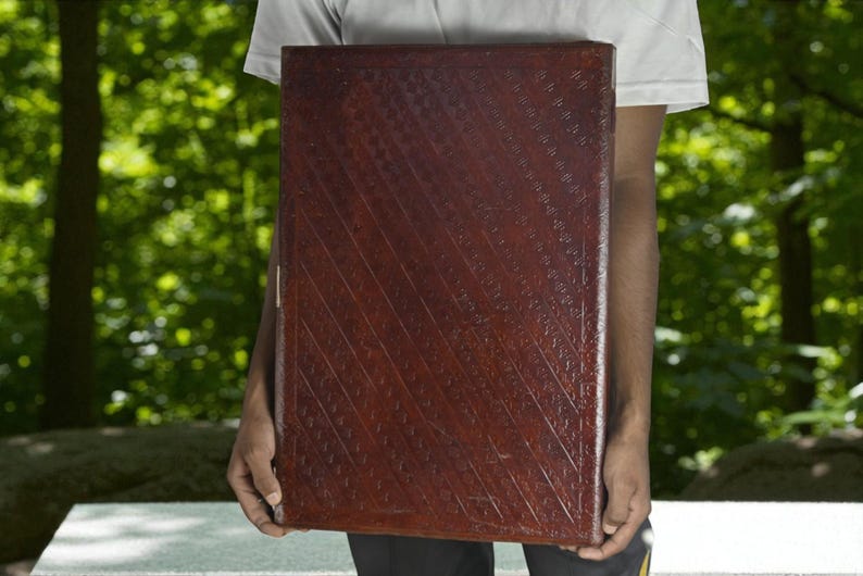 May include: A large, rectangular, dark brown leather-bound book with a textured, diagonal pattern. The book is held by a person, with a blurred green background of trees and foliage.