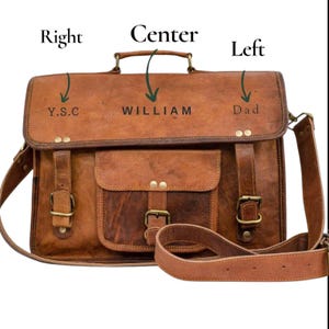 May include: A brown leather satchel with a shoulder strap and a front flap pocket. The bag has brass-colored buckles and the text "Y.S.C", "WILLIAM", and "Dad" embossed on the top. The bag has a handle and a detachable shoulder strap.