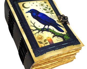 Può includere: Un diario rilegato in pelle con un'illustrazione di corvo. La copertina presenta un corvo blu scuro appollaiato su un ramo, su uno sfondo color crema con accenti floreali e celesti. Il diario ha una chiusura decorativa e pagine invecchiate.