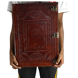 May include: A large, rectangular, brown leather journal with an embossed tree design and decorative border. The journal has a brass clasp closure and is held by a person. The leather has a rich, textured appearance.