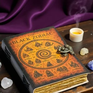 May include: A vintage-style leather book, titled "The Black Zodiac", with a decorative metal clasp. The cover is a mix of orange and black, featuring zodiac symbols and text. The aged pages are visible, with a candle and crystals.