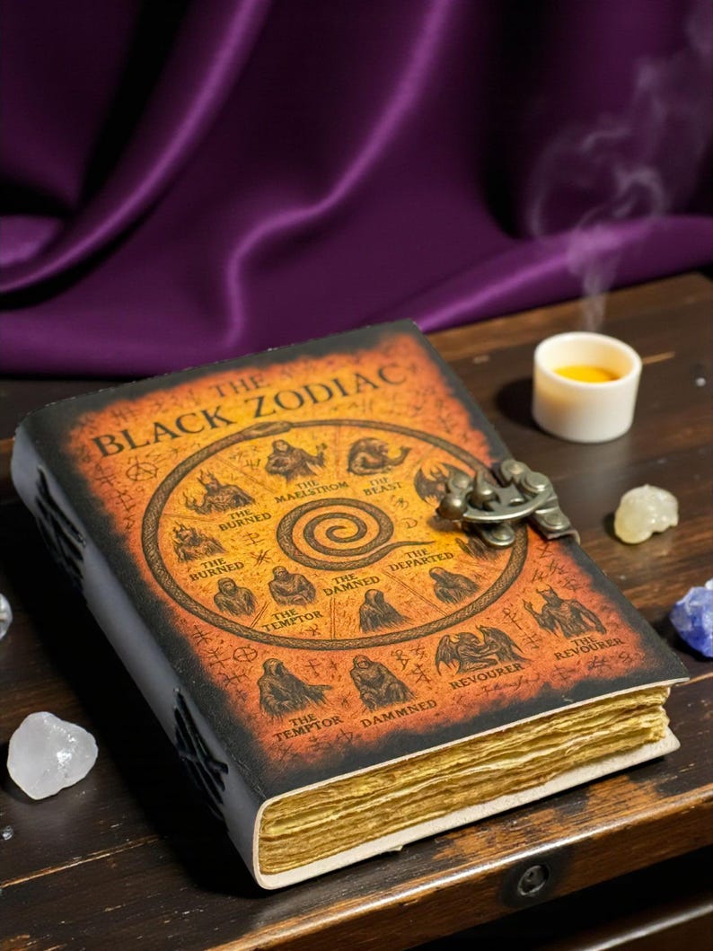 May include: A vintage-style leather book, titled "The Black Zodiac", with a decorative metal clasp. The cover is a mix of orange and black, featuring zodiac symbols and text. The aged pages are visible, with a candle and crystals.