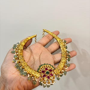 May include: A gold necklace with detailed floral engravings and a central floral pendant. The pendant features red and green gemstones, surrounded by small white beads and green teardrop-shaped stones. The necklace is held in a hand.