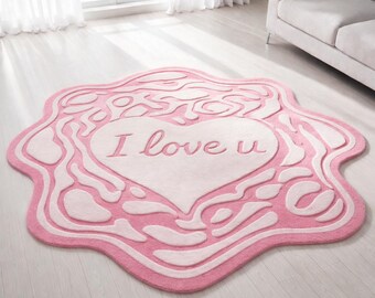 May include: Pink and white rug with a heart shape and the words "I love u" in the center. The rug has a unique, wavy border design and is placed on a light-colored wooden floor.