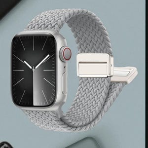 May include: A silver smartwatch with a gray braided band. The watch face is black and white with silver hands. The band has a silver clasp. The watch is displayed against a light blue background.