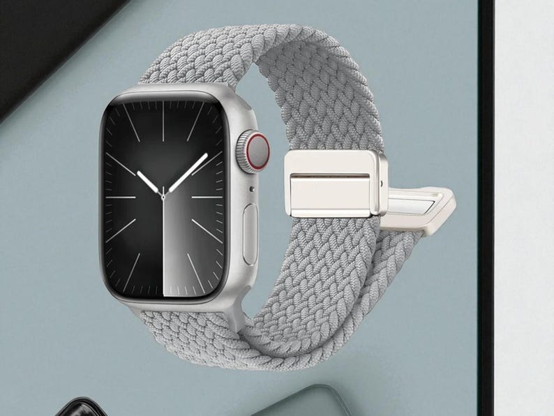 May include: A silver smartwatch with a gray braided band. The watch face is black and white with silver hands. The band has a silver clasp. The watch is displayed against a light blue background.