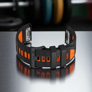 Puede incluir: Correa de reloj negra y naranja con hebilla. La correa presenta detalles rectangulares naranjas en los laterales y en la hebilla. Fabricada con material flexible, está diseñada para un smartwatch. Medidas en centímetros.